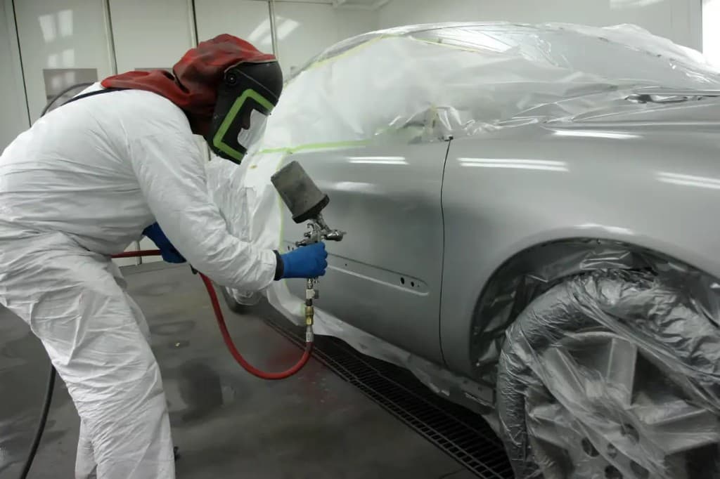 Painter in protective gear respraying a masked silver car in a spray booth